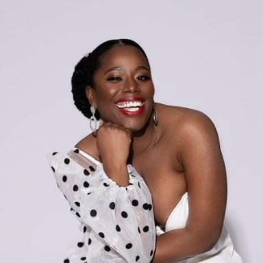 a black woman in a one-shoulder white dress, resting her chin on her loose fist and smiling into the camera