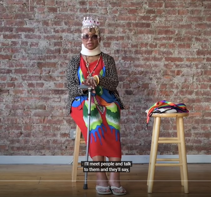 Rev. Goddess Kennedy a Black gender nonconforming sitting in front of a brick wall in a crown, bright dress, and sweater.